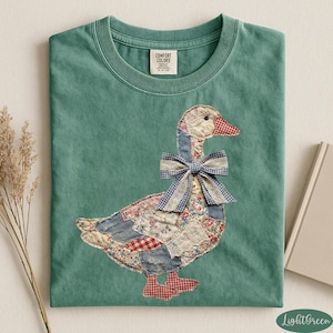 Peut inclure: T-shirt vert clair avec une application d'oie. L'oie est faite de tissu patchwork avec un n&oelig;ud en vichy bleu. Le t-shirt a un col rond et la marque "Comfort Colors" est visible sur l'&eacute;tiquette.