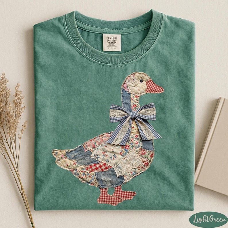 Peut inclure: T-shirt vert clair avec une application d'oie. L'oie est faite de tissu patchwork avec un n&oelig;ud en vichy bleu. Le t-shirt a un col rond et la marque "Comfort Colors" est visible sur l'&eacute;tiquette.