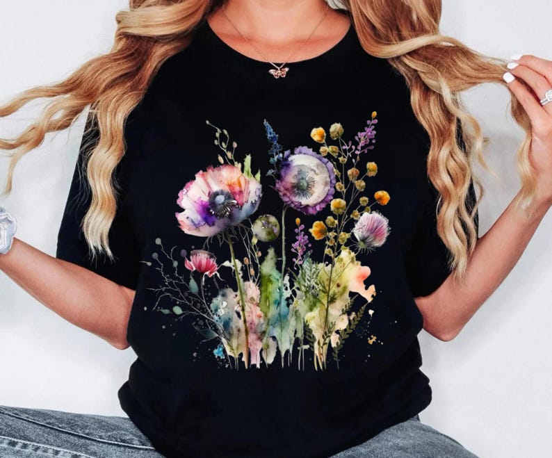 May include: Black t-shirt featuring a watercolor floral design. The design includes pink, purple, yellow, and green flowers and foliage. The flowers are the focal point, with detailed petals and leaves.