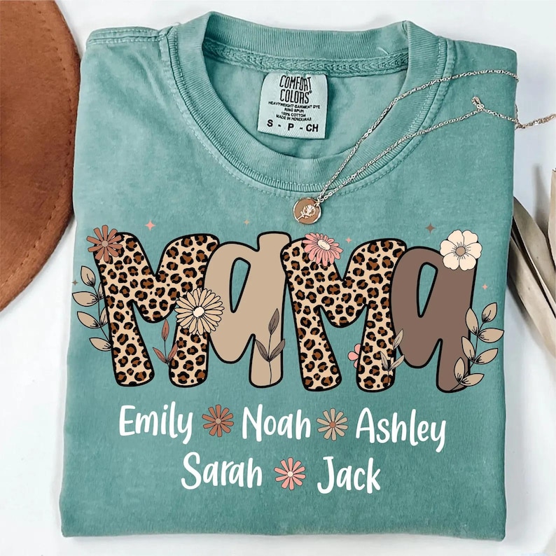 May include: A teal Comfort Colors t-shirt with the word "MAMA" in a mix of leopard print and tan letters, decorated with floral accents. Below the word are the names Emily, Noah, Ashley, Sarah, and Jack.