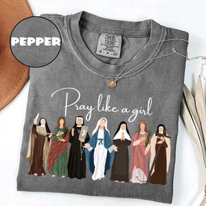 May include: A gray Comfort Colors t-shirt with the text "Pray like a girl" and an illustration of seven women. The shirt has a crew neck and short sleeves. A silver necklace with a pendant is on the shirt.