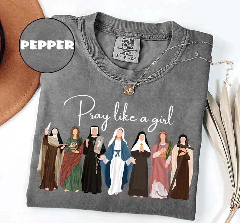 May include: A gray Comfort Colors t-shirt with the text "Pray like a girl" and an illustration of seven women. The shirt has a crew neck and short sleeves. A silver necklace with a pendant is on the shirt.