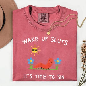 Puede incluir: Una camiseta roja con el texto "WAKE UP SLUTS IT'S TIME TO SIN" y un dibujo de un gusano sonriente, un sol y flores. Un collar de estrella dorada est&aacute; sobre la camiseta. La camiseta es talla XL-TG-EG.