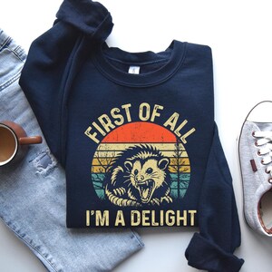 May include: Navy blue sweatshirt with a retro graphic of an opossum inside a circle with the text "FIRST OF ALL I'M A DELIGHT". The design features orange, yellow, and green stripes, and a vintage aesthetic.