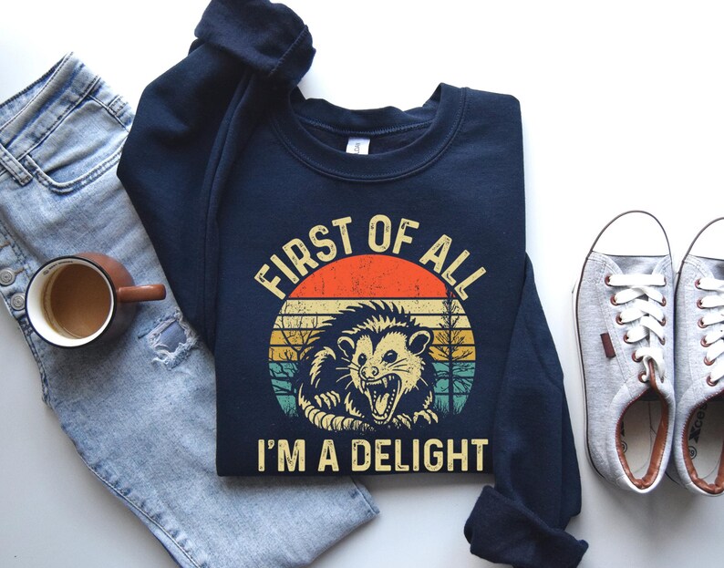 May include: Navy blue sweatshirt with a retro graphic of an opossum inside a circle with the text "FIRST OF ALL I'M A DELIGHT". The design features orange, yellow, and green stripes, and a vintage aesthetic.