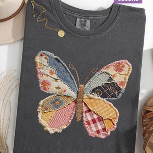 May include: A dark gray t-shirt with a large, patchwork butterfly design. The butterfly wings feature various fabric patterns in shades of blue, pink, yellow, and red. A gold necklace with a pendant is draped on the shirt. The text "COMFORT COLORS" is visible.