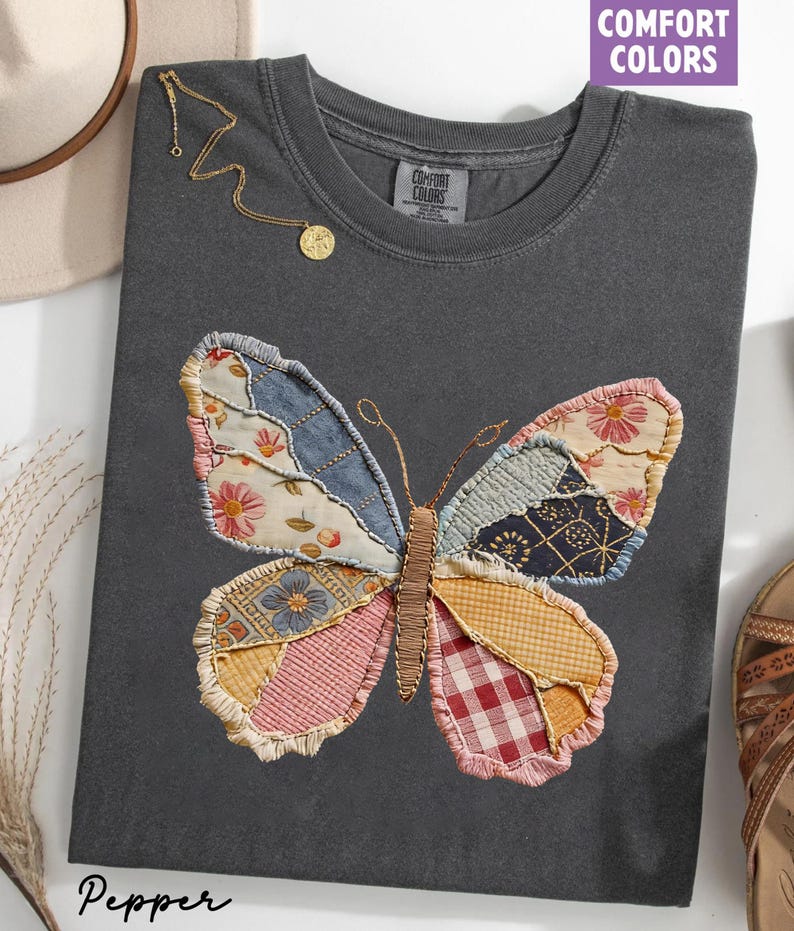 May include: A dark gray t-shirt with a large, patchwork butterfly design. The butterfly wings feature various fabric patterns in shades of blue, pink, yellow, and red. A gold necklace with a pendant is draped on the shirt. The text "COMFORT COLORS" is visible.