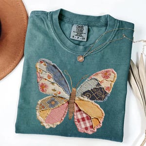 May include: Teal Comfort Colors t-shirt featuring a patchwork butterfly design. The butterfly wings are made of various patterned fabrics in shades of blue, pink, yellow, and red. A gold-colored necklace with a pendant rests on the shirt. The tag reads "Comfort Colors".