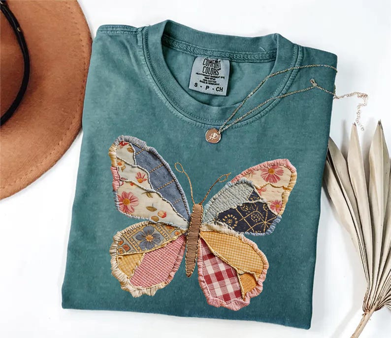 May include: Teal Comfort Colors t-shirt featuring a patchwork butterfly design. The butterfly wings are made of various patterned fabrics in shades of blue, pink, yellow, and red. A gold-colored necklace with a pendant rests on the shirt. The tag reads "Comfort Colors".
