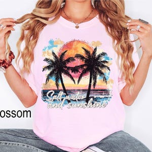 Op de afbeelding: Roze T-shirt met een vintage-stijl grafische afbeelding van palmbomen en een zonsondergang. De tekst "Salt water and sunshine" is op het shirt gedrukt.