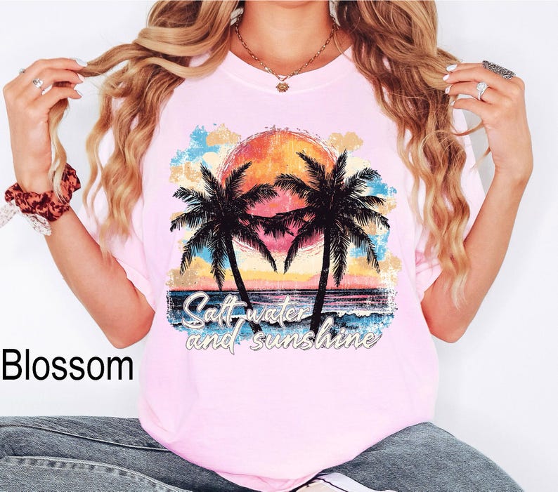 Op de afbeelding: Roze T-shirt met een vintage-stijl grafische afbeelding van palmbomen en een zonsondergang. De tekst "Salt water and sunshine" is op het shirt gedrukt.