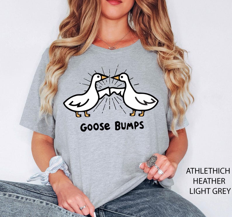 May include: Light grey t-shirt featuring a cartoon design of two white geese with black outlines and yellow feet, facing each other. The text "Goose Bumps" is printed below the geese in black. The shirt also has the text "ATHLETHICH HEATHER LIGHT GREY".