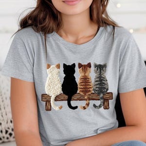 May include: A light grey t-shirt with an embroidered design of four cats on a wooden bench. The cats are white, black, brown, and grey, seen from the back. The t-shirt is made of a soft, comfortable material.