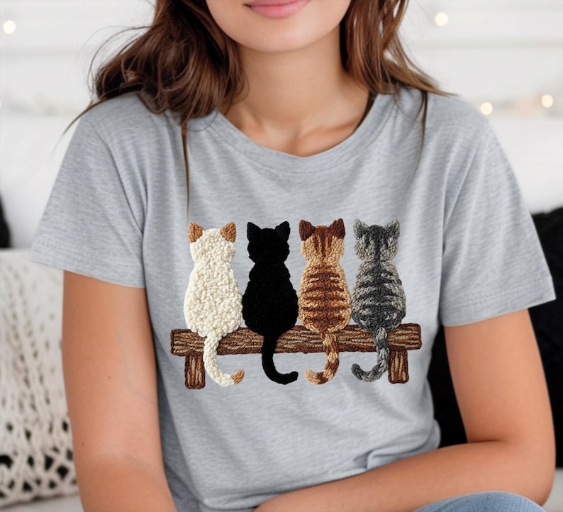 May include: A light grey t-shirt with an embroidered design of four cats on a wooden bench. The cats are white, black, brown, and grey, seen from the back. The t-shirt is made of a soft, comfortable material.