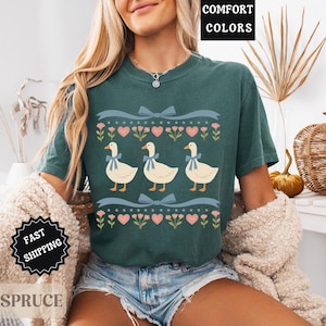 Pode incluir: Uma camiseta verde-azulado com um design fantasioso de tr&ecirc;s gansos com la&ccedil;os azuis, em um fundo de cora&ccedil;&otilde;es e padr&otilde;es florais. A camisa tem o texto "Comfort Colors" em uma caixa preta.