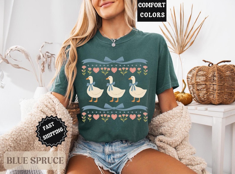 Pode incluir: Uma camiseta verde-azulado com um design fantasioso de tr&ecirc;s gansos com la&ccedil;os azuis, em um fundo de cora&ccedil;&otilde;es e padr&otilde;es florais. A camisa tem o texto "Comfort Colors" em uma caixa preta.