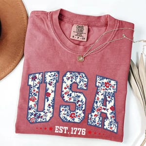 May include: A dusty rose t-shirt with the word "USA" in a floral pattern. The shirt also features the text "EST. 1776" in white. A gold necklace and a brown hat are visible in the image.