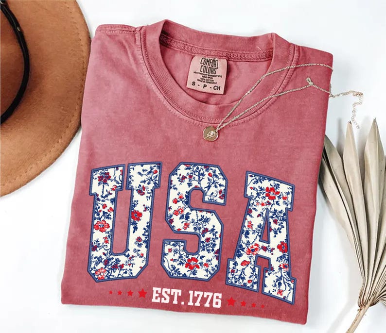 May include: A dusty rose t-shirt with the word "USA" in a floral pattern. The shirt also features the text "EST. 1776" in white. A gold necklace and a brown hat are visible in the image.