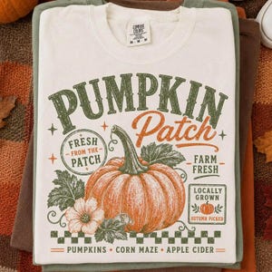 May include: A cream-colored t-shirt featuring a graphic print with the text PUMPKIN Patch, FRESH FROM THE PATCH, FARM FRESH, LOCALLY GROWN, AUTUMN PICKED, PUMPKINS, CORN MAZE, and APPLE CIDER. The design includes a large orange pumpkin and leaves.