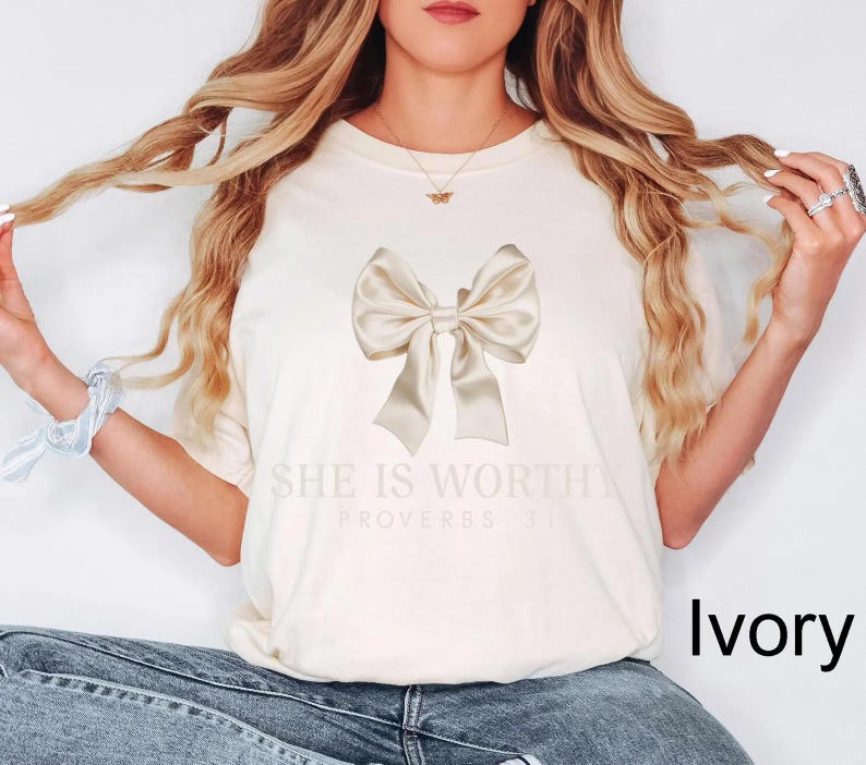 May include: Ivory t-shirt with a large beige satin bow and the text "She is Worthy Proverbs 31:25"