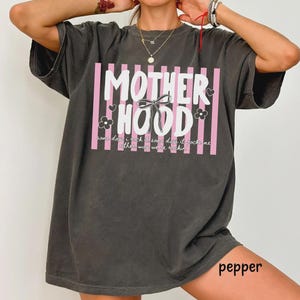 May include: Dark gray t-shirt with the text "MOTHERHOOD" in white, set against pink vertical stripes. The design includes a bow, hearts, and flowers. The shirt also has the text "some days I rock, some days it rocks me, either way we're rockin".