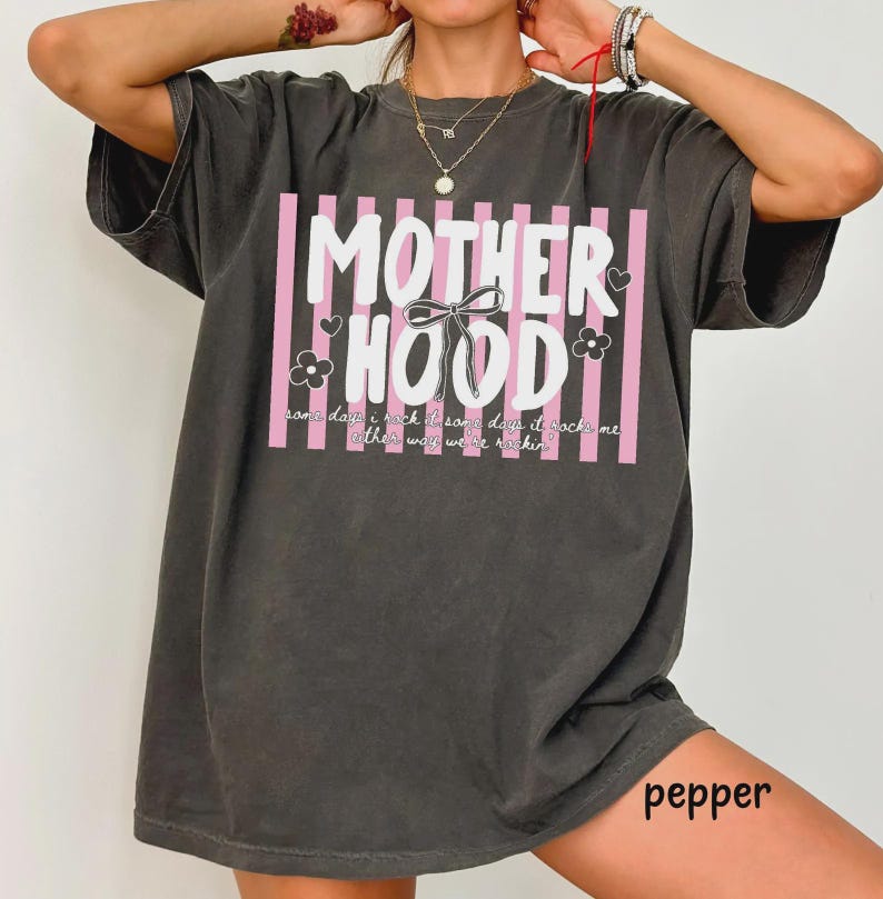 May include: Dark gray t-shirt with the text "MOTHERHOOD" in white, set against pink vertical stripes. The design includes a bow, hearts, and flowers. The shirt also has the text "some days I rock, some days it rocks me, either way we're rockin".