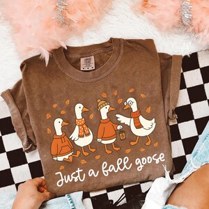 Puede incluir: Camiseta marr&oacute;n con una ilustraci&oacute;n de cuatro gansos vestidos con ropa de oto&ntilde;o, incluyendo bufandas y sombreros. El texto "Just a fall goose" est&aacute; escrito debajo. La camiseta tiene un corte holgado y es de material suave.