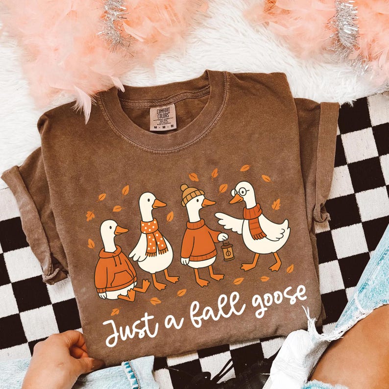 Puede incluir: Camiseta marr&oacute;n con una ilustraci&oacute;n de cuatro gansos vestidos con ropa de oto&ntilde;o, incluyendo bufandas y sombreros. El texto "Just a fall goose" est&aacute; escrito debajo. La camiseta tiene un corte holgado y es de material suave.