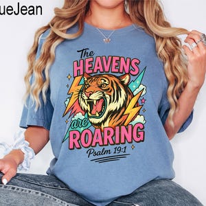 Puede incluir: Camiseta azul claro con el texto "The Heavens are Roaring" y un gr&aacute;fico de un tigre rugiendo. El dise&ntilde;o incluye rayos y el vers&iacute;culo b&iacute;blico Salmo 19:1.