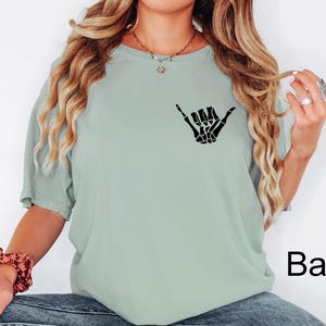 Pode incluir: Uma camiseta verde claro com uma m&atilde;o de esqueleto preta fazendo um sinal shaka. O texto "Bay" &eacute; vis&iacute;vel no canto inferior direito da imagem.