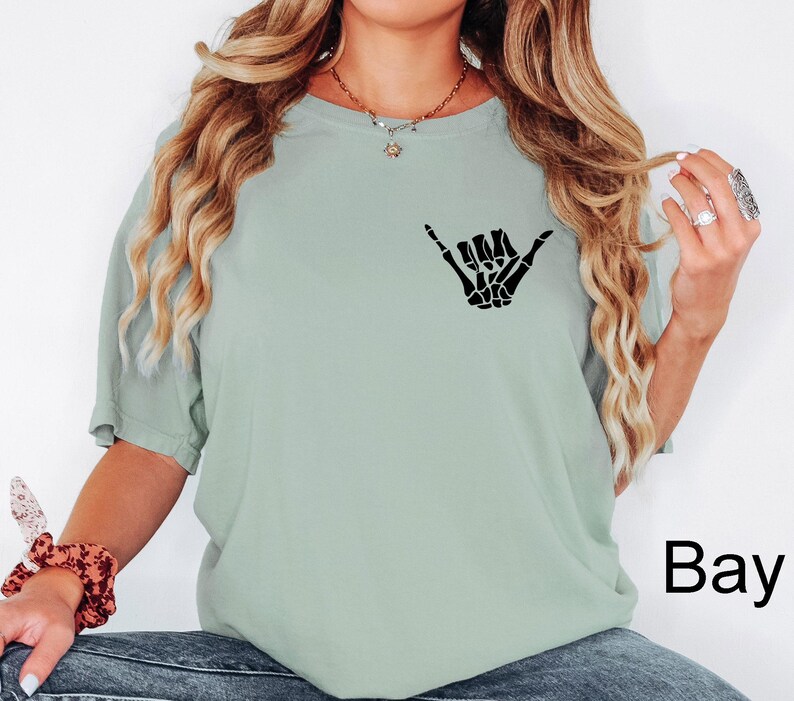 Pode incluir: Uma camiseta verde claro com uma m&atilde;o de esqueleto preta fazendo um sinal shaka. O texto "Bay" &eacute; vis&iacute;vel no canto inferior direito da imagem.