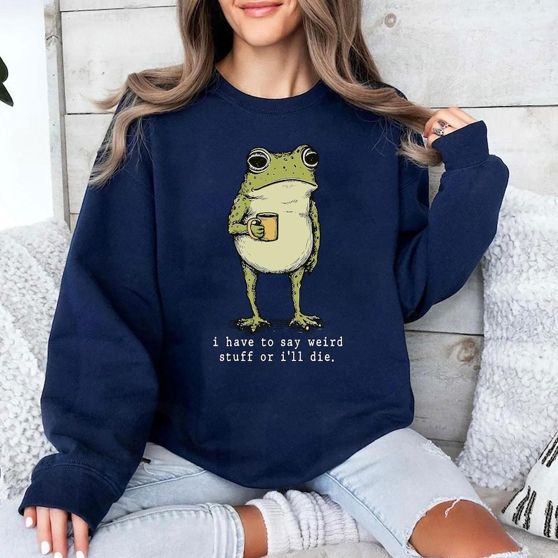 May include: Navy blue sweatshirt featuring a cartoon frog holding a mug. The frog is green and beige with large eyes. The text "I have to say weird stuff or I'll die" is printed below the frog in white. The sweatshirt is a casual style.