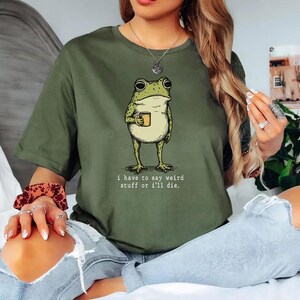 May include: Olive green t-shirt featuring a whimsical illustration of a frog holding a mug. The frog is light green with a cream-colored belly. The text "I have to say weird stuff or I'll die." is printed below the frog.