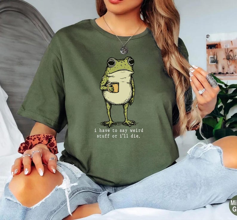 May include: Olive green t-shirt featuring a whimsical illustration of a frog holding a mug. The frog is light green with a cream-colored belly. The text "I have to say weird stuff or I'll die." is printed below the frog.