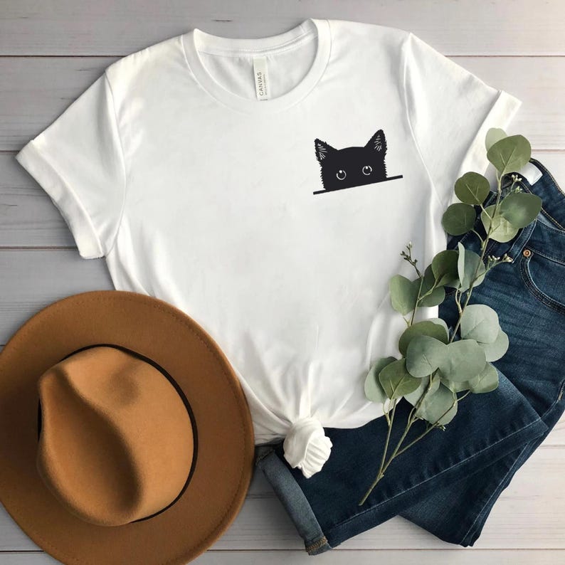 May include: A white short-sleeved t-shirt featuring a black graphic of a cat head peeking over a horizontal line on the left chest area. The shirt has a crew neckline and is shown with a tan hat, blue denim jeans, and a green eucalyptus sprig.