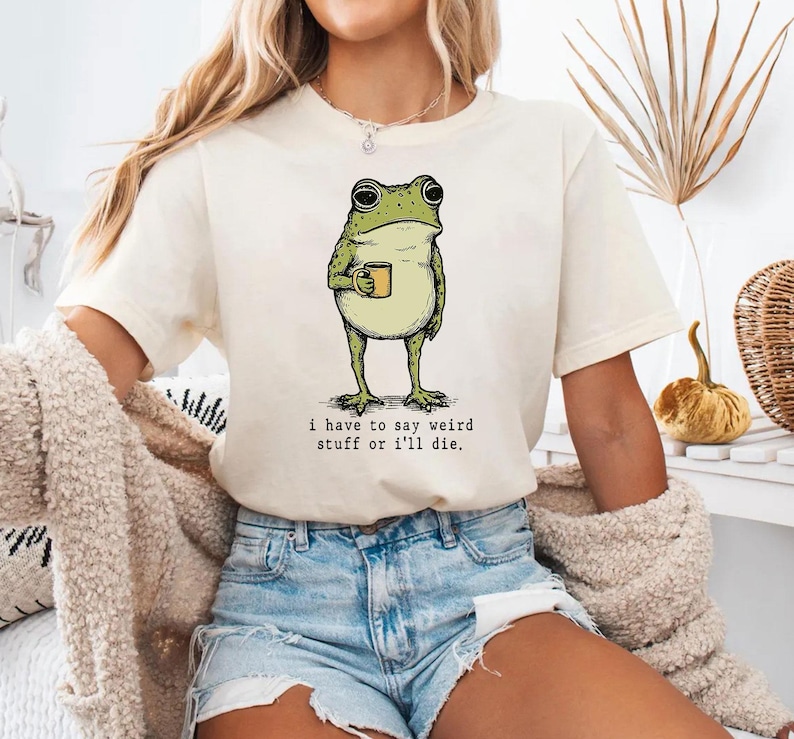 May include: Cream-colored t-shirt featuring a green frog holding a yellow mug. The frog illustration has a vintage style. The text "i have to say weird stuff or i'll die" is printed below the frog. The shirt is made of soft material.