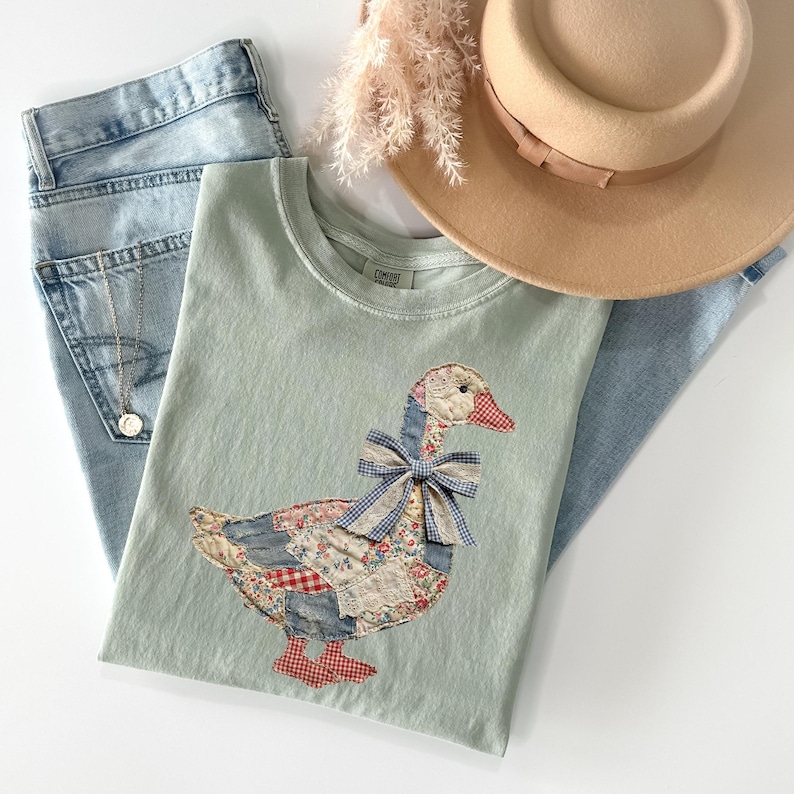Peut inclure: Un t-shirt vert clair avec un motif d'oie appliqu&eacute;, avec un corps en patchwork, un n&oelig;ud en vichy bleu et des pieds et un bec &agrave; carreaux rouges et blancs. Le t-shirt est associ&eacute; &agrave; un jean bleu clair et un chapeau beige.