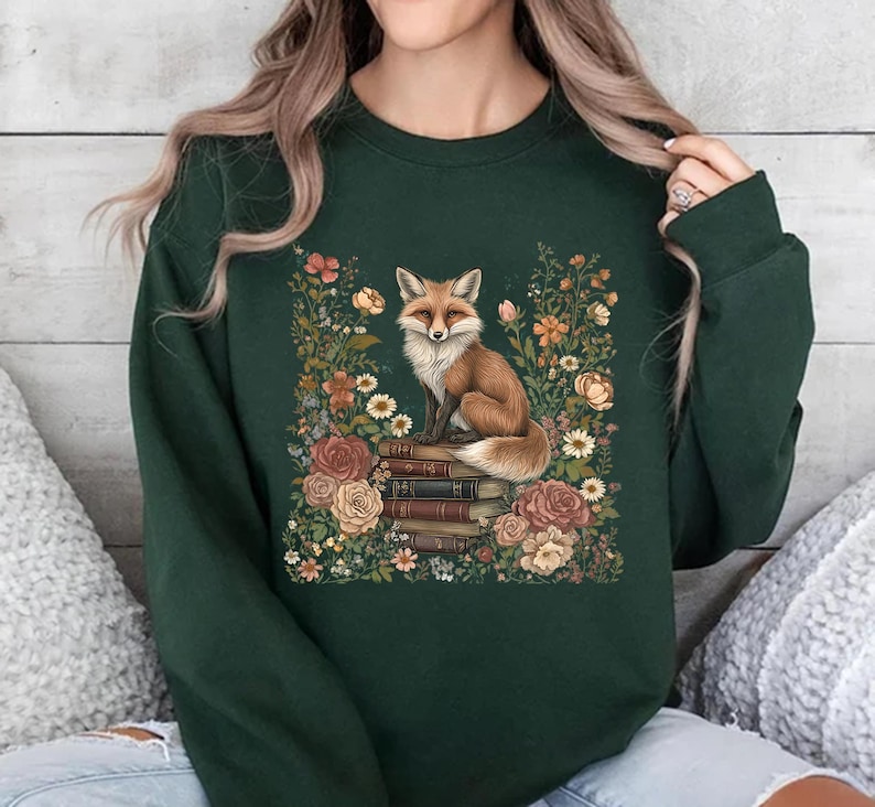 K&ouml;nnte beinhalten: Ein dunkelgr&uuml;nes Sweatshirt mit Rundhalsausschnitt und einem zentralen Grafikdruck eines Fuchses, der auf einem Stapel alter B&uuml;cher sitzt, umgeben von einem farbenfrohen Arrangement aus verschiedenen Blumen und Bl&auml;ttern in Orange-, Rosa- und Cremet&ouml;nen.