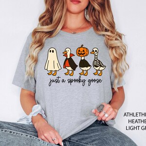 以下が含まれることがあります： ハロウィーンをテーマにしたデザインのライトグレーのTシャツ。グラフィックには、幽霊、吸血鬼のマントを着たガチョウ、カボチャの頭のガチョウ、スケルトンのガチョウが含まれています。「just a spooky goose」というテキストが下にあります。