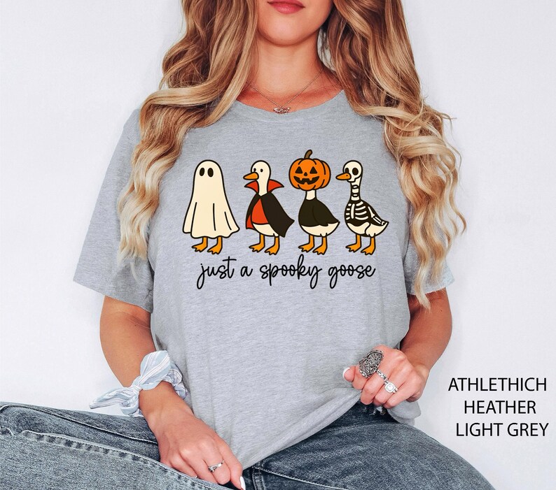 以下が含まれることがあります： ハロウィーンをテーマにしたデザインのライトグレーのTシャツ。グラフィックには、幽霊、吸血鬼のマントを着たガチョウ、カボチャの頭のガチョウ、スケルトンのガチョウが含まれています。「just a spooky goose」というテキストが下にあります。
