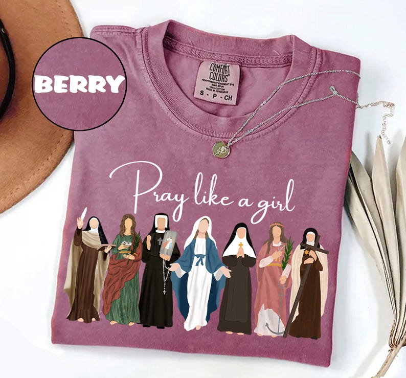 May include: Berry-colored t-shirt with the phrase "Pray like a girl" in cursive. The shirt features an illustration of seven women in religious attire. A silver necklace with a pendant is draped across the shirt.