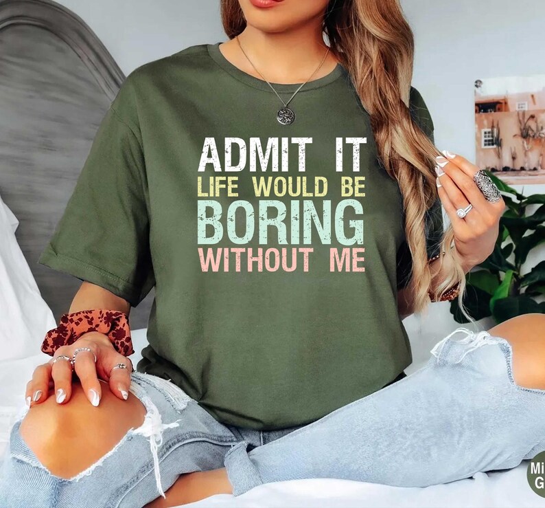 May include: Olive green t-shirt with the text "ADMIT IT LIFE WOULD BE BORING WITHOUT ME" in white, light green, and pink. The shirt is a casual, crew neck style.
