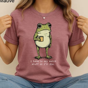 May include: Mauve t-shirt featuring a cartoon frog holding a mug. The frog is green and beige with large eyes. The text "i have to say weird stuff or i'll die" is printed below the frog in a simple font.