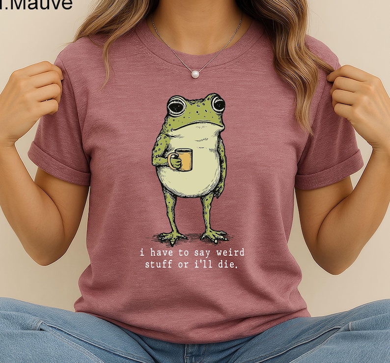 May include: Mauve t-shirt featuring a cartoon frog holding a mug. The frog is green and beige with large eyes. The text "i have to say weird stuff or i'll die" is printed below the frog in a simple font.