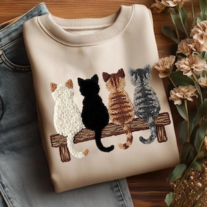 May include: A cream-coloured sweatshirt with an embroidered design of four cats on a wooden bench. The cats are white, black, brown, and grey. The sweatshirt is next to blue jeans and flowers.