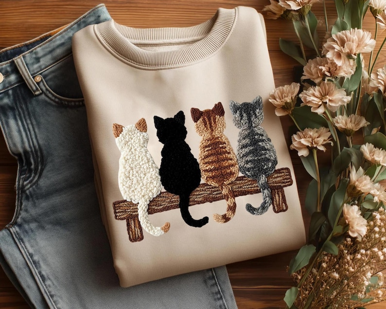 May include: A cream-coloured sweatshirt with an embroidered design of four cats on a wooden bench. The cats are white, black, brown, and grey. The sweatshirt is next to blue jeans and flowers.
