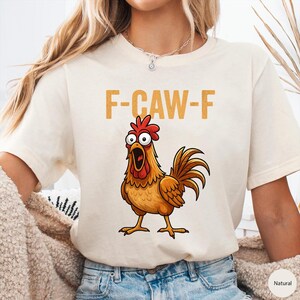 May include: A natural-colored short-sleeve t-shirt featuring a graphic of a wide-eyed, orange-brown rooster with an open beak. Above the rooster, the text F-CAW-F is printed in a bold, mustard-yellow font. The shirt has a crew neckline.