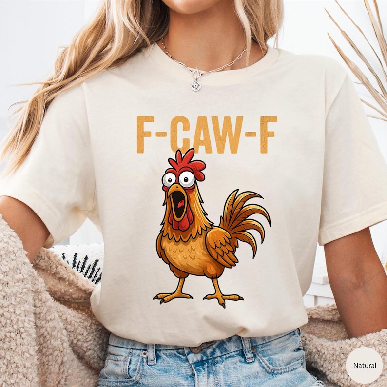 May include: A natural-colored short-sleeve t-shirt featuring a graphic of a wide-eyed, orange-brown rooster with an open beak. Above the rooster, the text F-CAW-F is printed in a bold, mustard-yellow font. The shirt has a crew neckline.