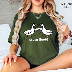 May include: Military green t-shirt featuring a graphic of two white geese facing each other with a sunburst design between them. The text "Goose Bumps" is below the geese in white. The shirt is a solid color.