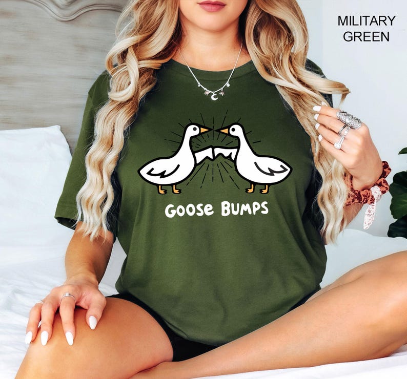 May include: Military green t-shirt featuring a graphic of two white geese facing each other with a sunburst design between them. The text "Goose Bumps" is below the geese in white. The shirt is a solid color.